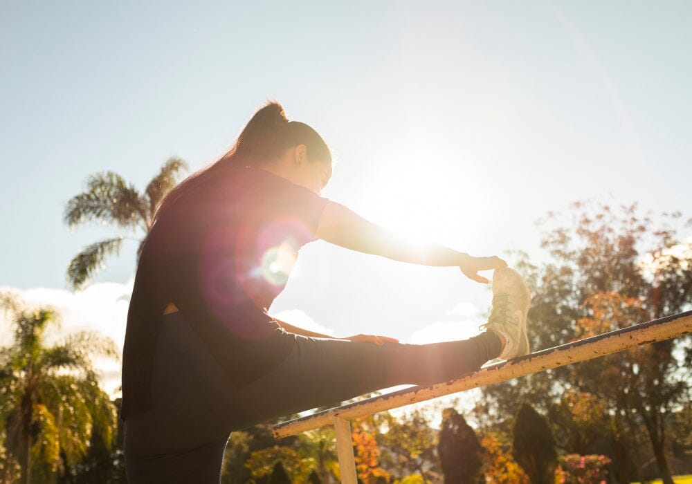 A woman stretching her leg on a railing outside with the bright sun behind her 