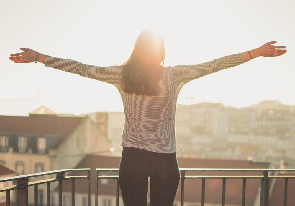 A woman standing on a balcony facing the sun with her arms outstretched