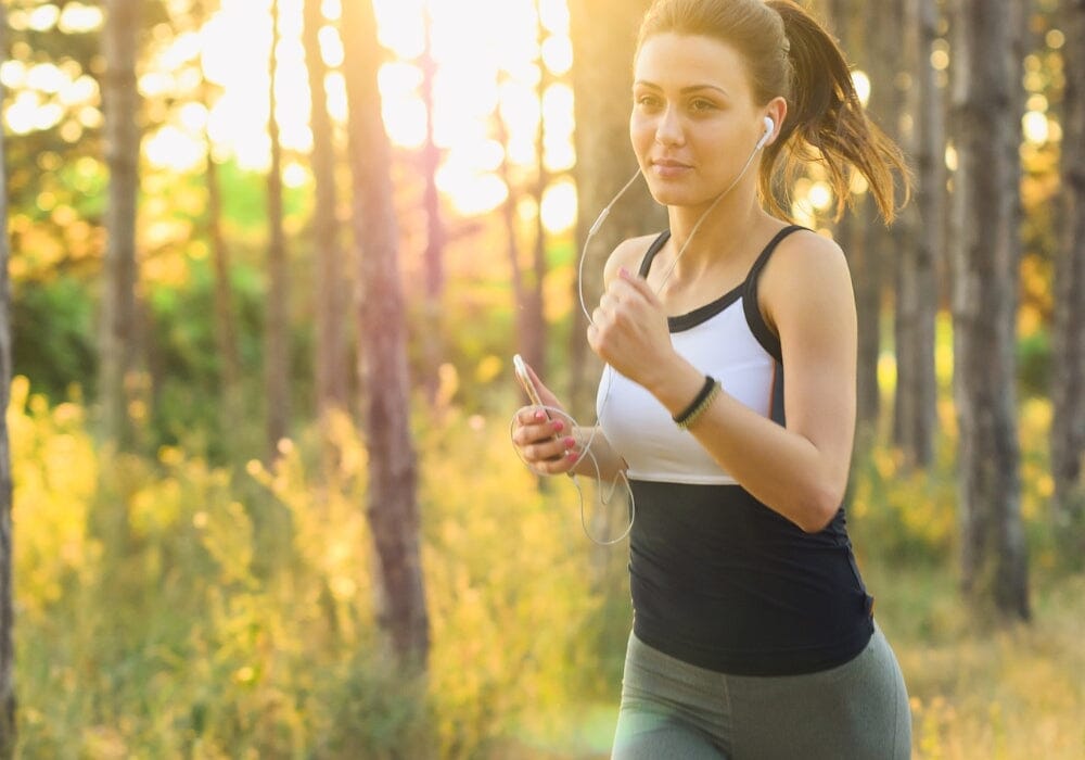 A woman running in the woods wearing earphones and holding a smartphone