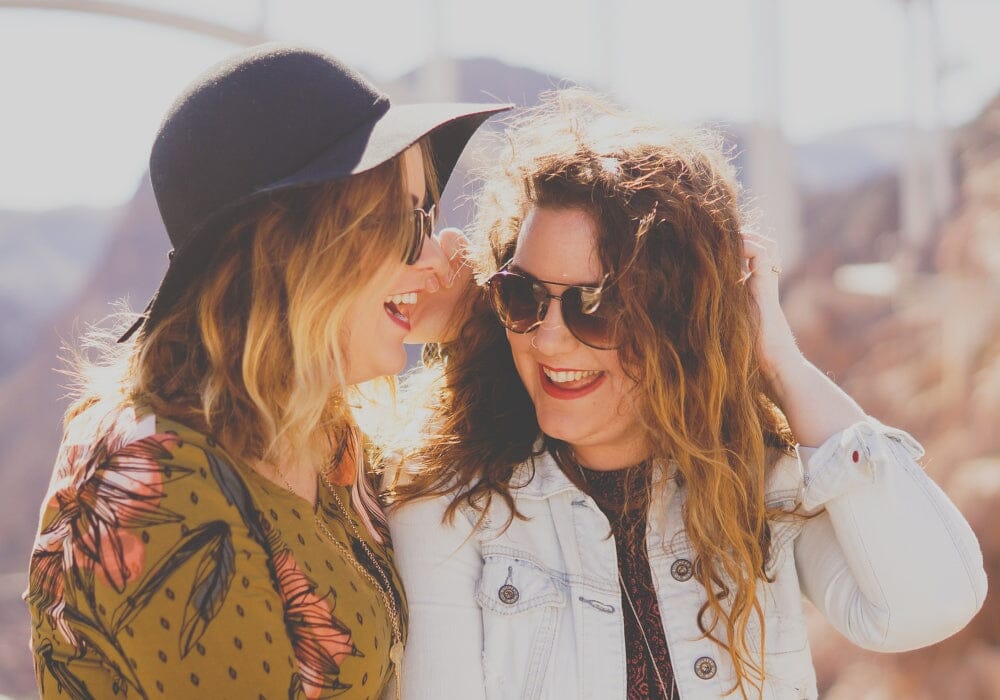 Two women, both in sunglasses, one wearing a hat, talking and laughing together