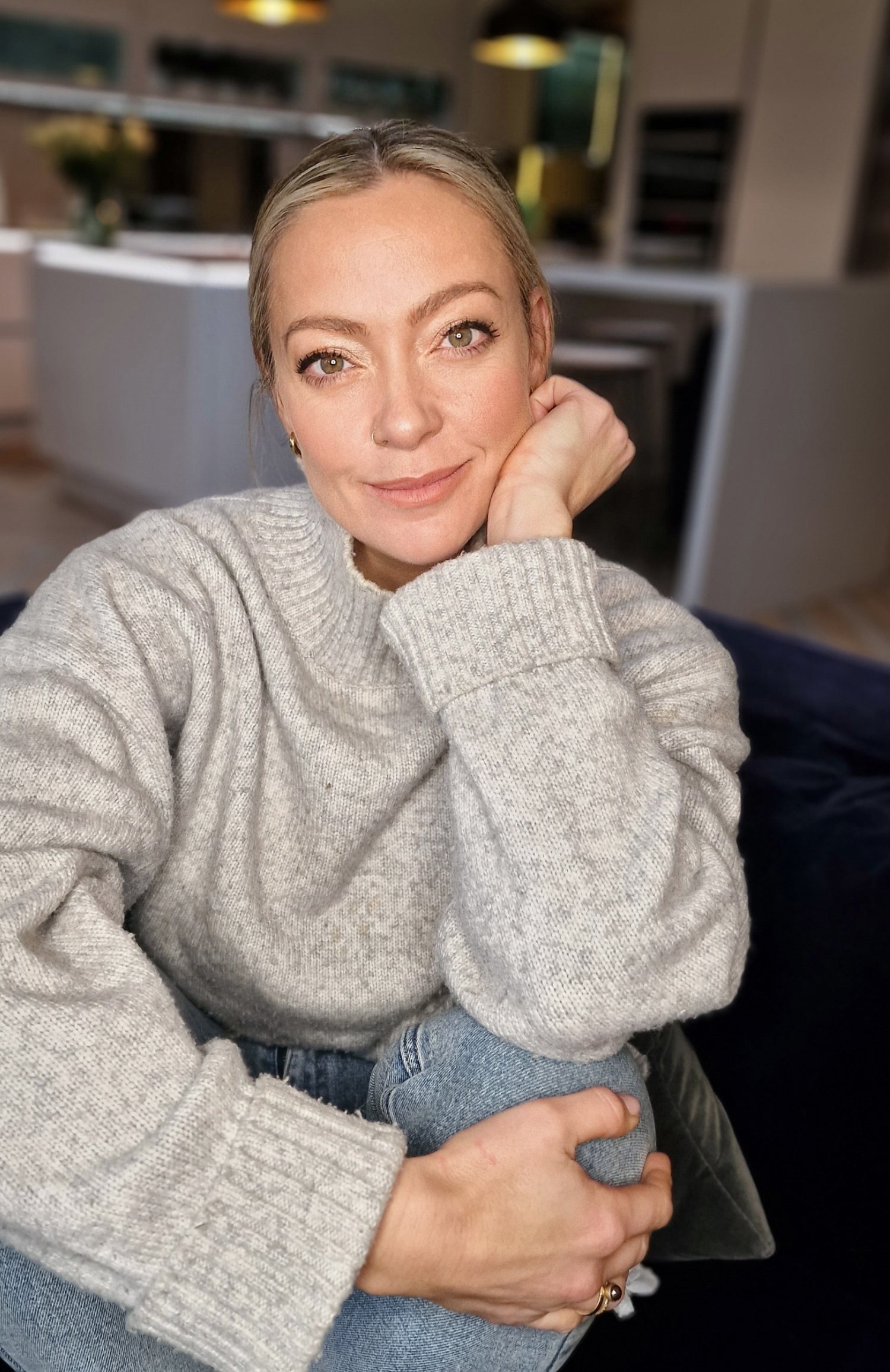 Woman smiling, sitting on sofa in grey jumper.