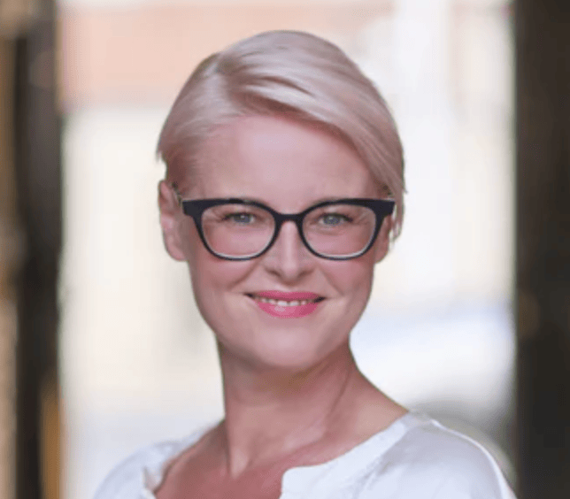 Woman with short blonde hair and glasses smiling against a blurred background